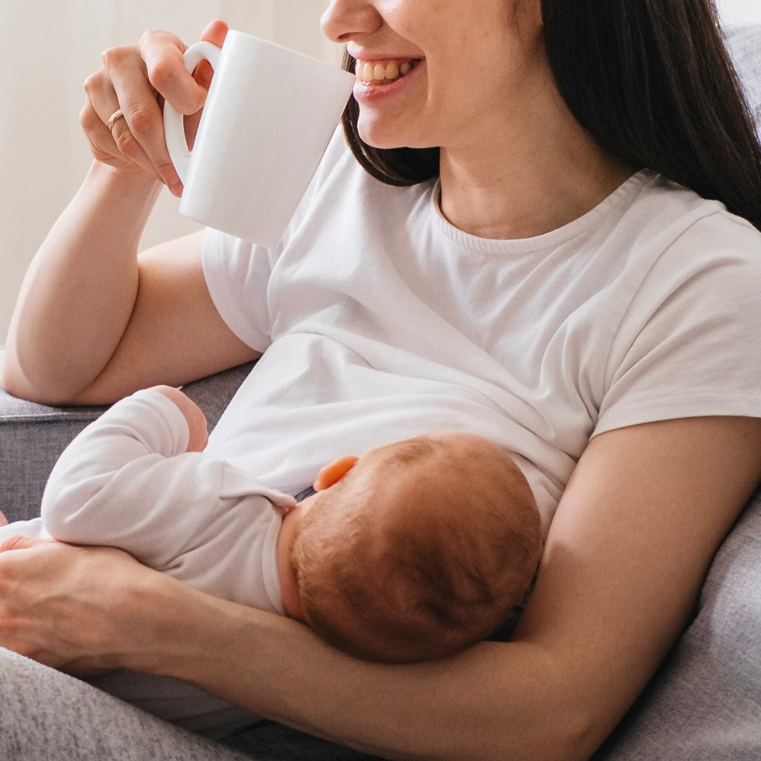 A woman in a white t-shirt is smiling while holding a mug and sitting in a comfortable chair. She is cradling a sleeping baby in her arms, who is dressed in light clothing. The scene conveys a sense of warmth and relaxation.